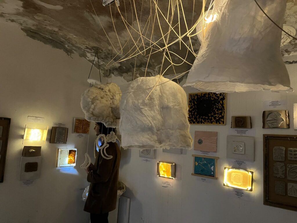 Exhibition room with lit boxes and framed works under suspended fabric forms